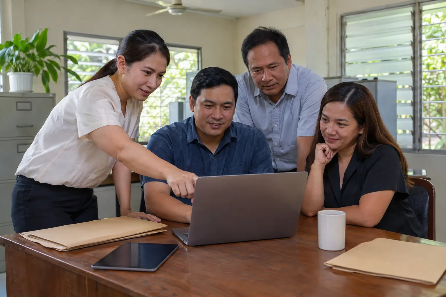 Illustrative: office staff gathered around a laptop while one colleague points at the screen